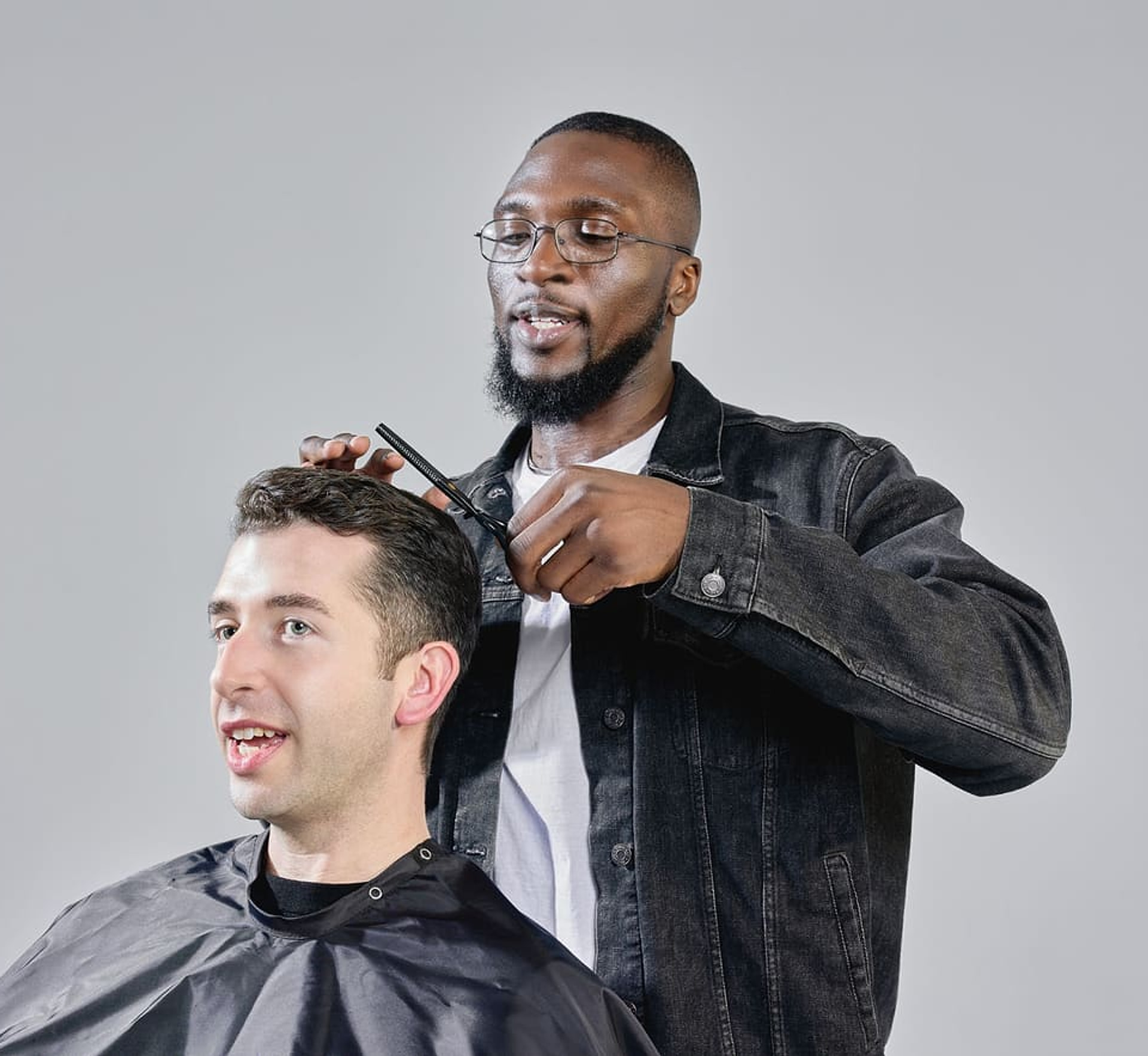 A barber trimming a seated client’s hair with scissors against a gray studio backdrop