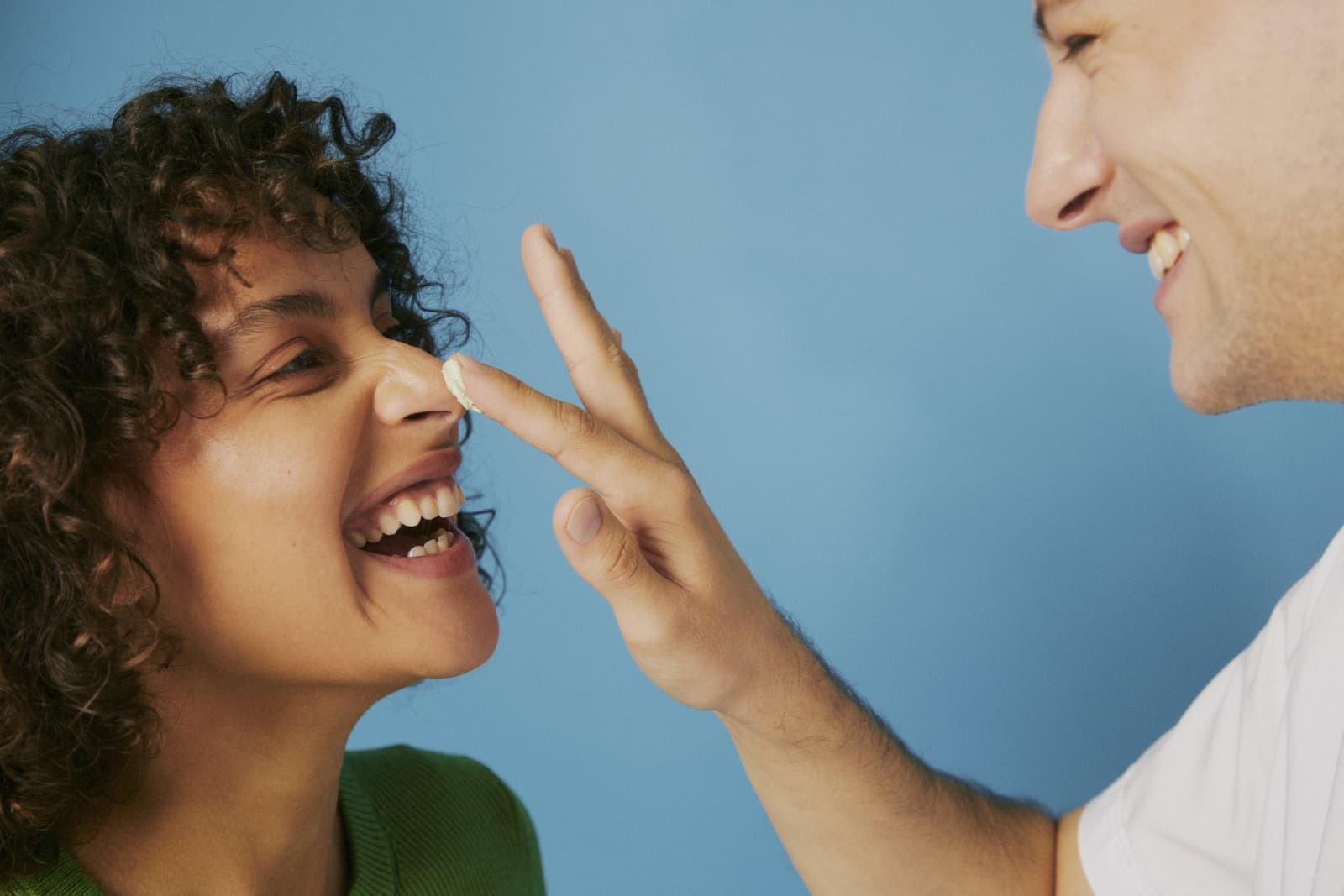Two people laughing against a blue backdrop as one dabs a dot of facial cream on the other’s nose