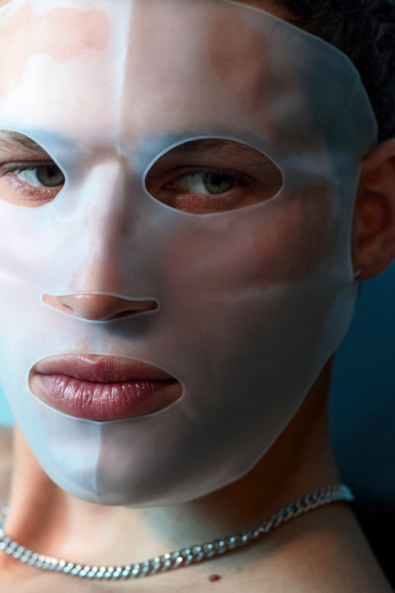 A close-up portrait of a person wearing a translucent frosted silicone face mask against a blue backdrop