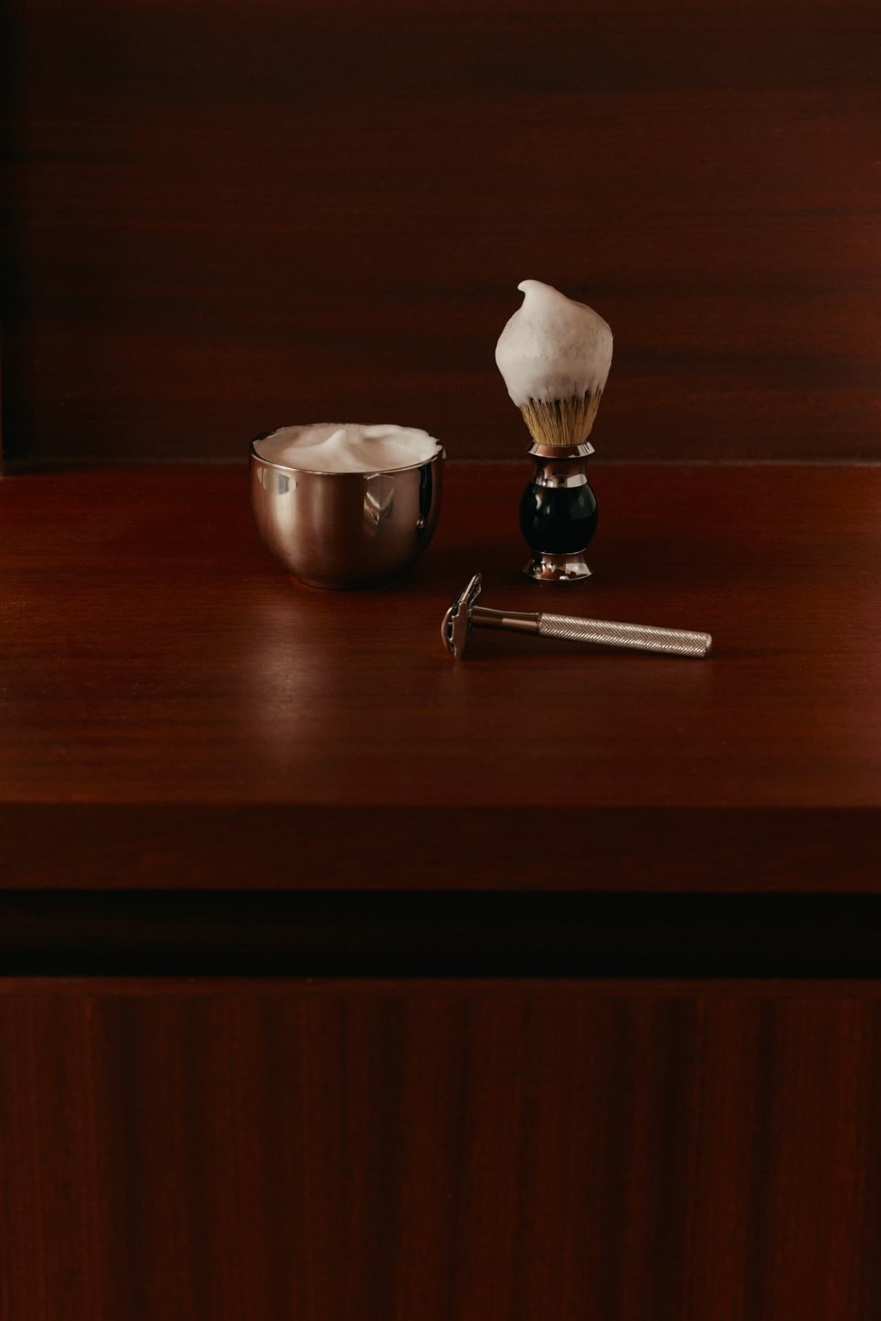 A traditional wet-shaving station on a dark mahogany counter — a polished steel lather bowl, a lathered shaving brush, and a knurled safety razor