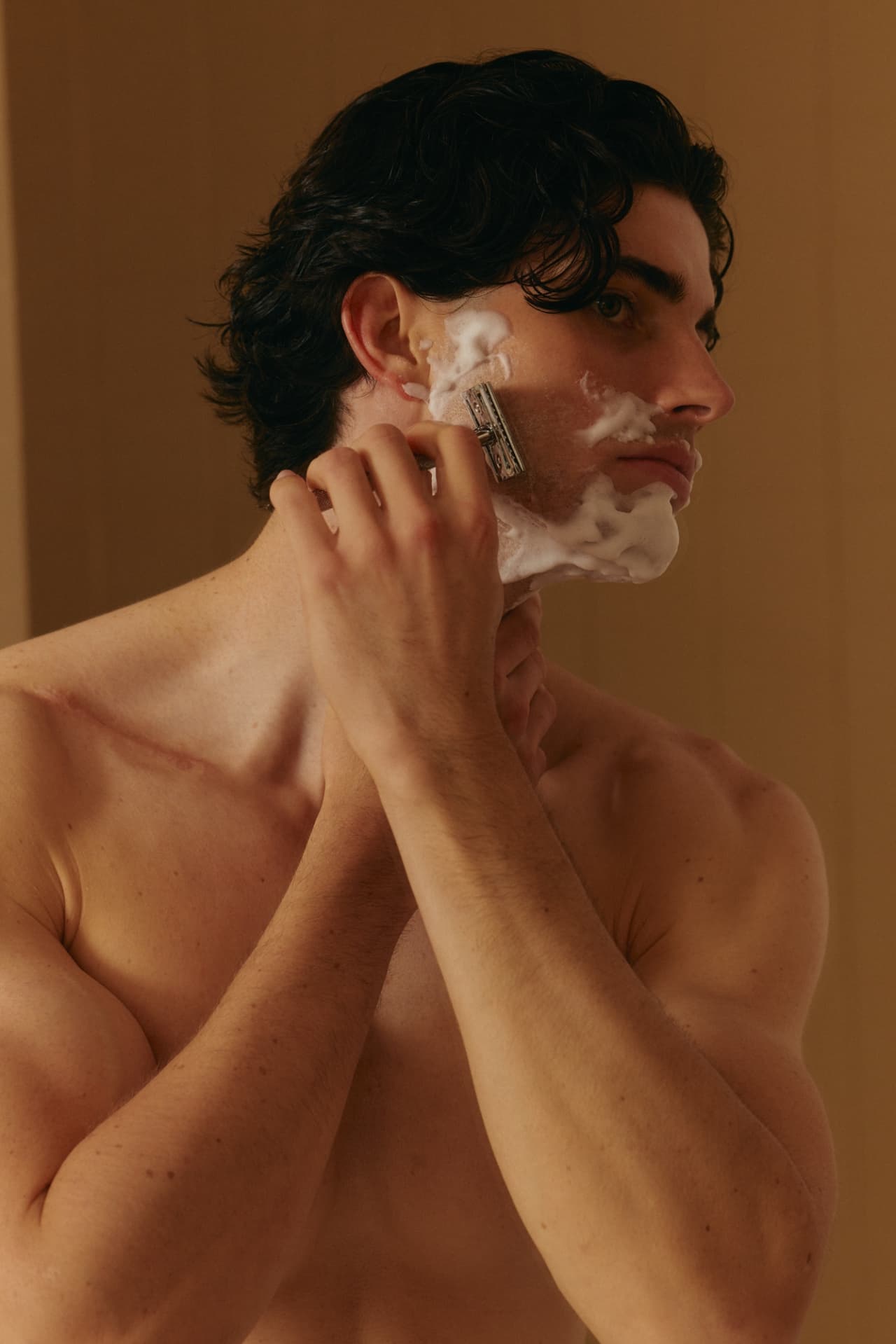 A man with dark hair shaving his face with a safety razor, his cheek covered in lather