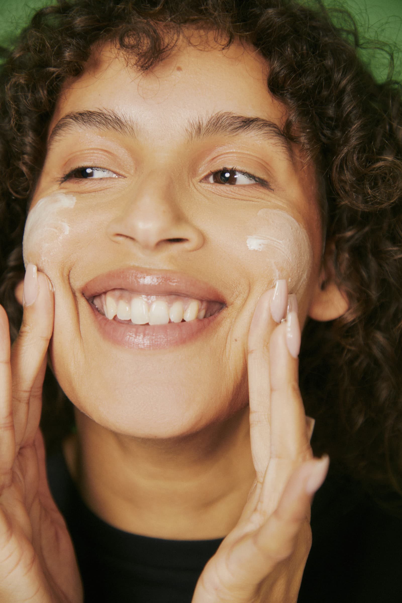A smiling client with curly hair applying cream to her cheeks against a green backdrop
