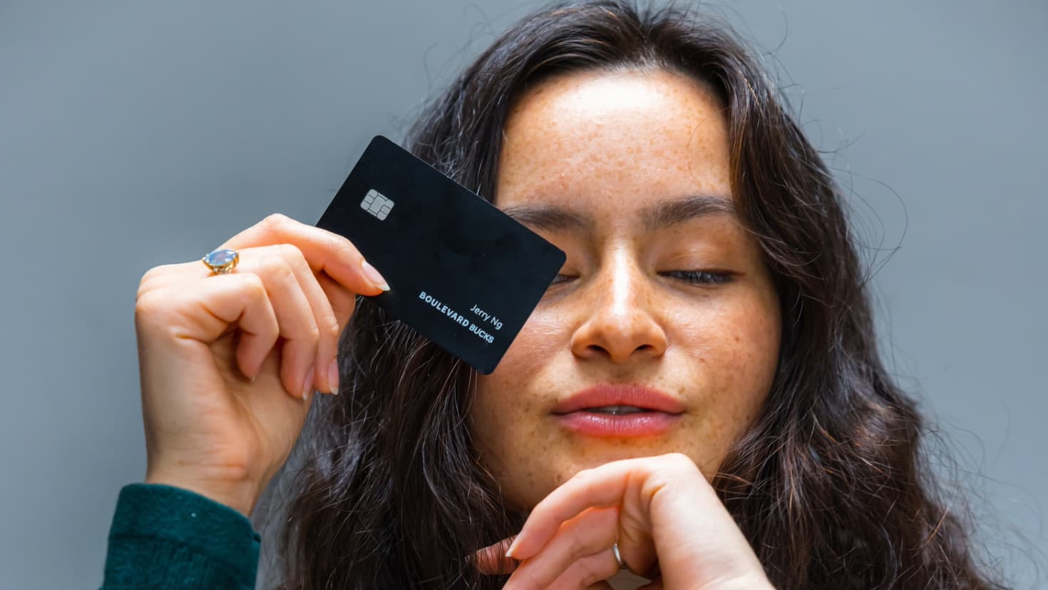 A person with long wavy hair has one hand on her chin and the other is holding a black credit card. 
