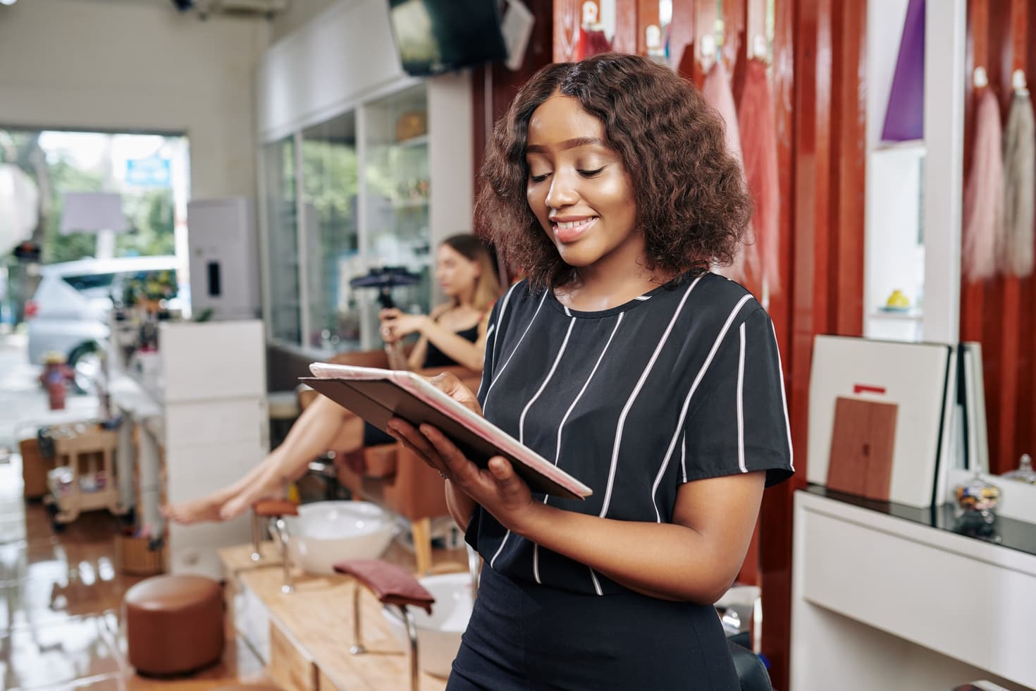 A smiling woman at a beauty salon checks her tablet while a client waits for a pedicure in the background
