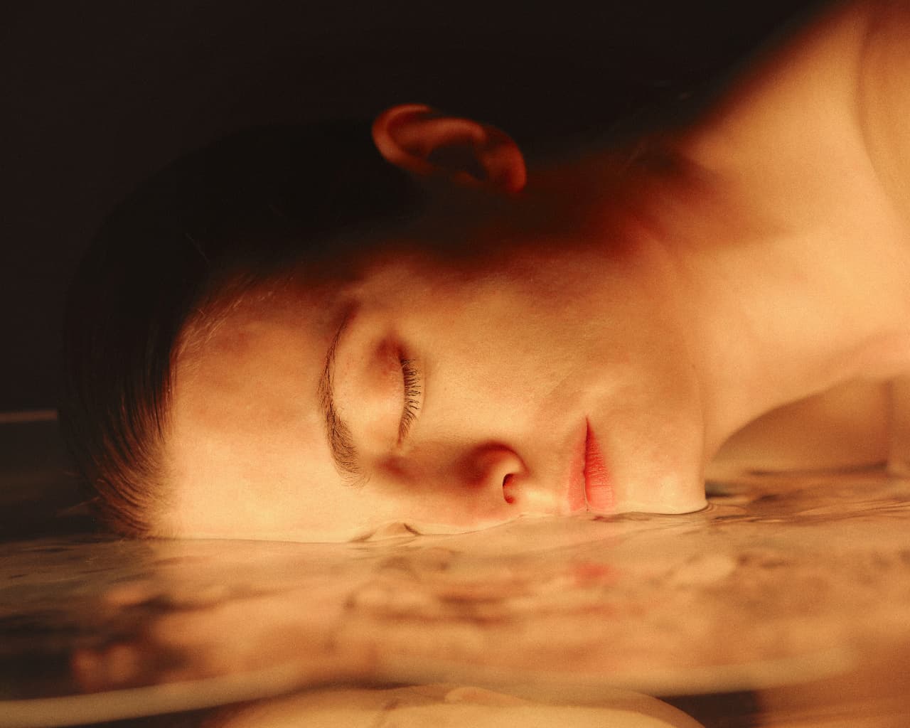 A person resting their cheek on the surface of still, warmly lit water with their eyes closed
