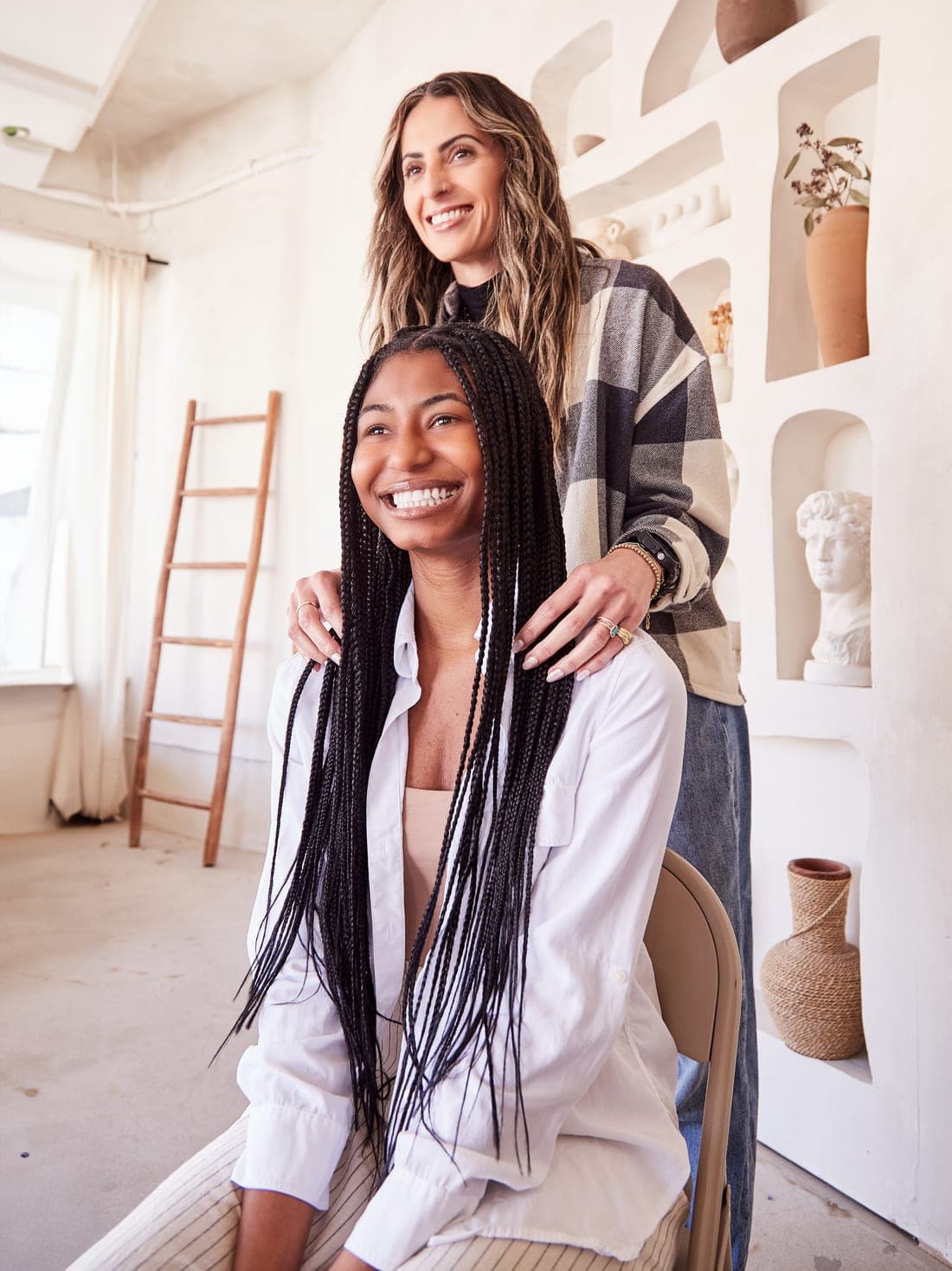 A smiling salon client with long braids, her stylist standing behind her with hands on her shoulders
