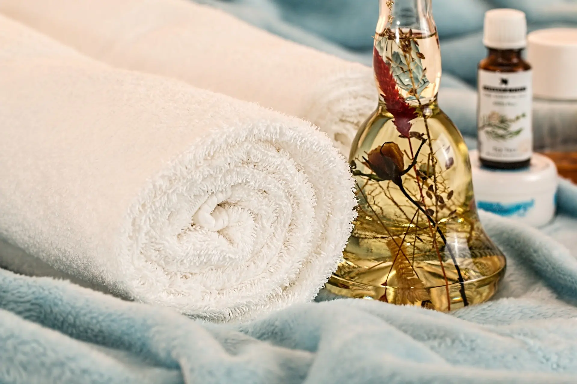 Rolled white towels on a soft blue blanket beside a glass bottle of botanical oil and small skincare jars
