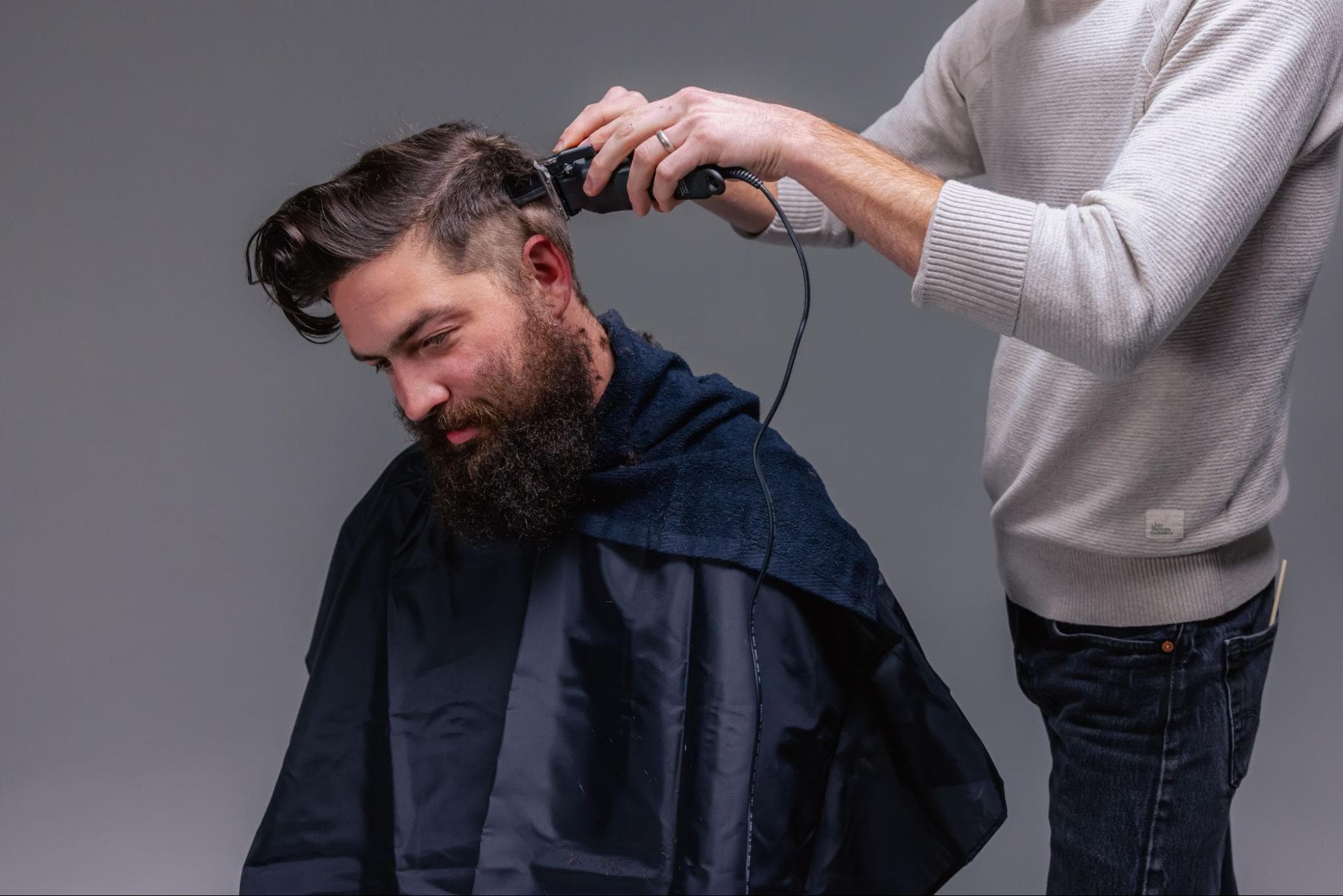 A seated person in a hair-cutting cape bends forward to allow another person, whose face is off frame, to clip their hair.