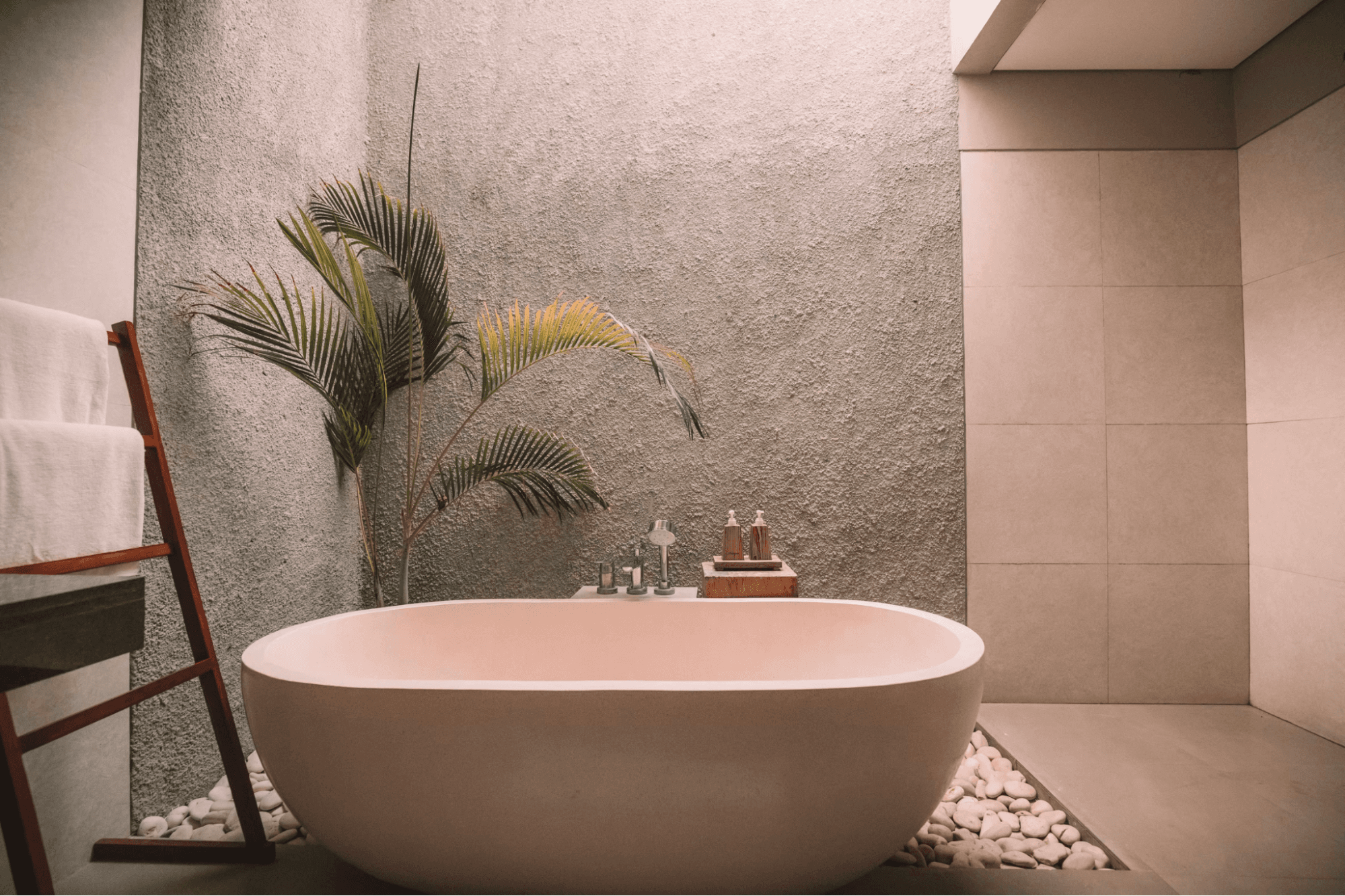 A serene spa treatment room with a freestanding soaking tub, folded towels, river stones, and a potted palm against a textured stone wall