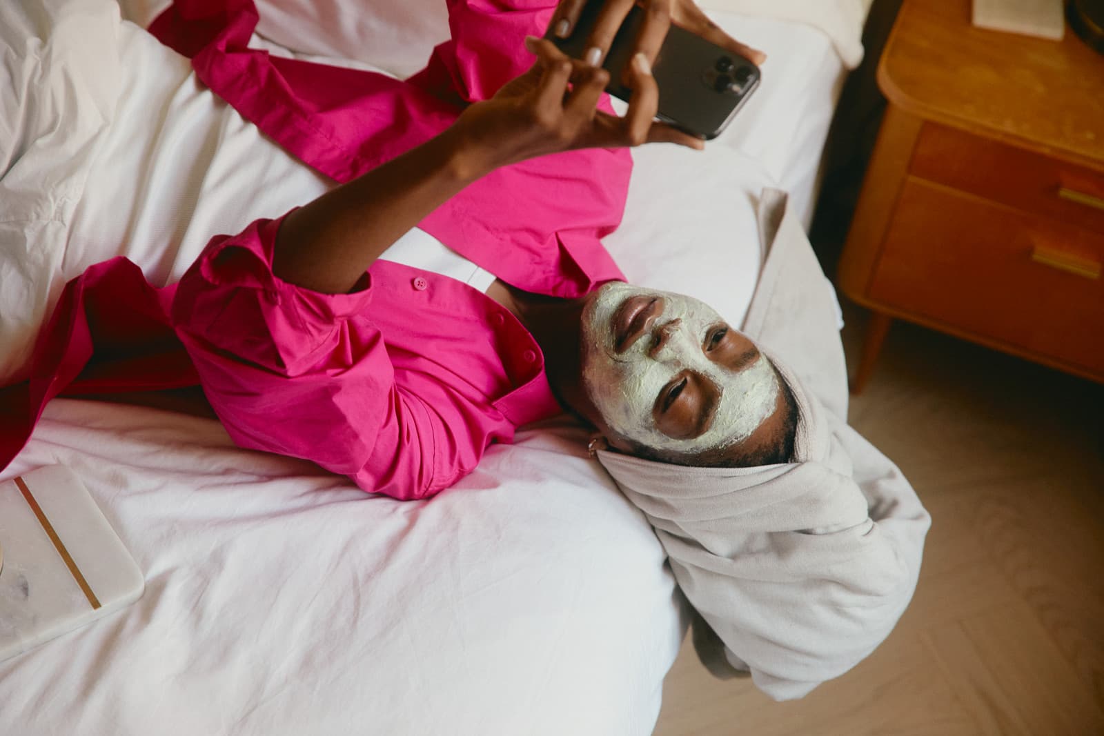 A client wearing a face mask lies in bed checking their phone, reflecting the self-service, on-your-own-schedule side of client booking