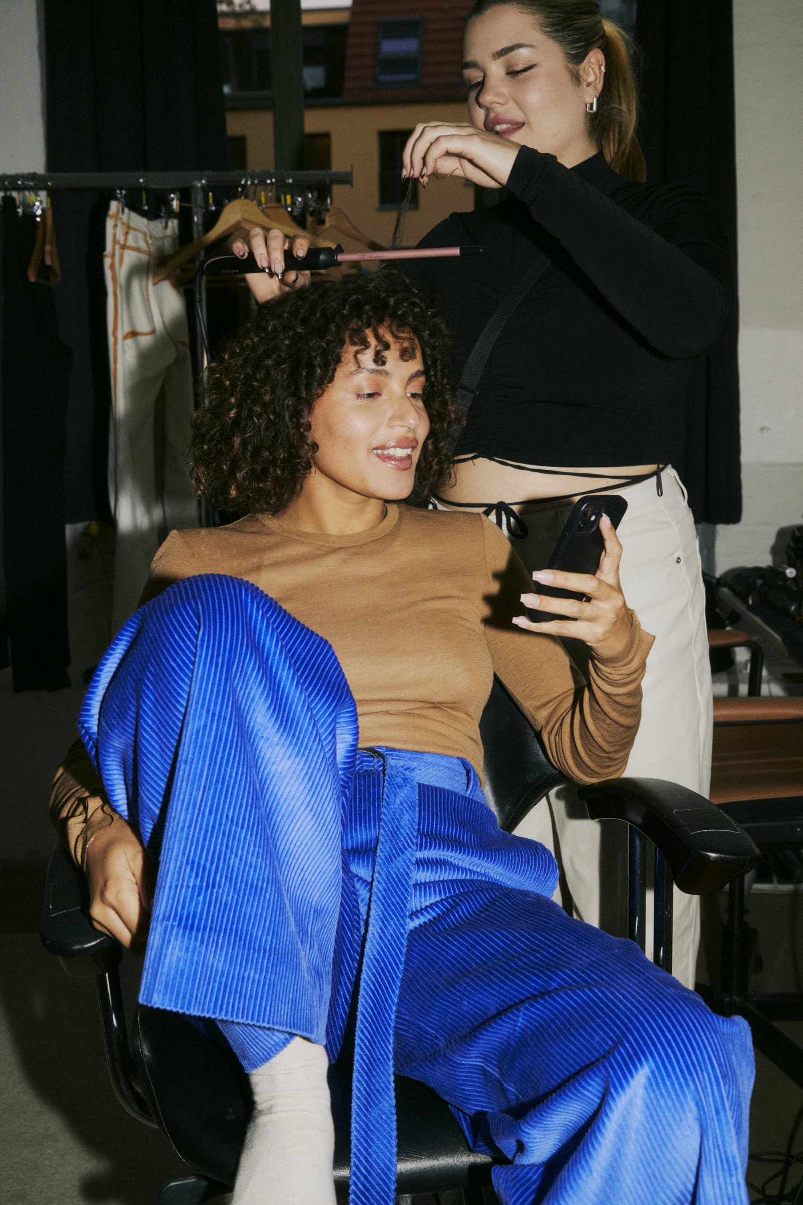 A hair stylist curls a smiling client’s hair with a curling iron while the client, seated in a salon chair, looks at her phone.