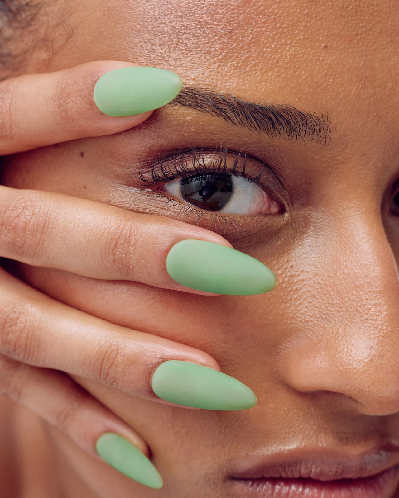 Close-up of a woman covering part of her face with her hand, showing long almond-shaped nails painted in matte sage green.