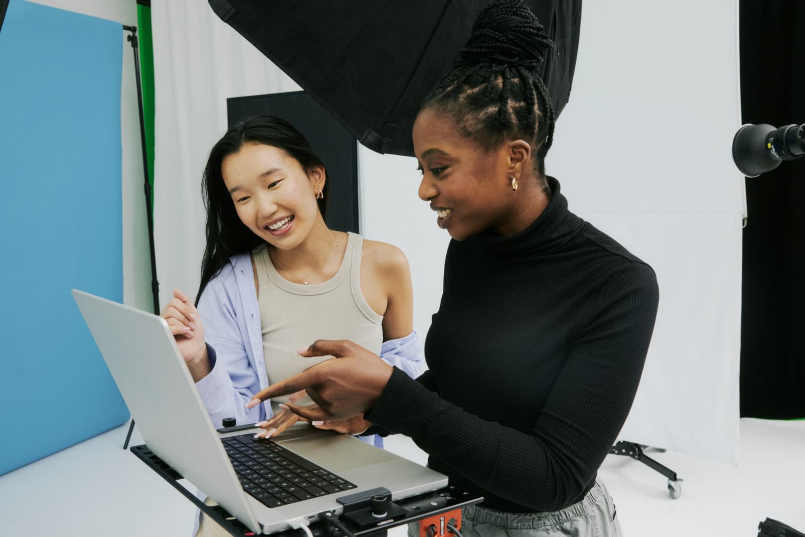 Two smiling women review content together on a laptop in a photo studio with lighting equipment and a blue backdrop