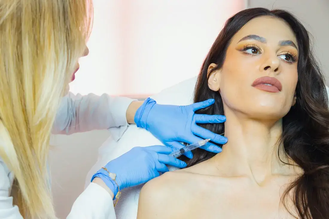 An aesthetics provider wearing blue gloves administers an injectable treatment to a client's neck in a bright treatment room