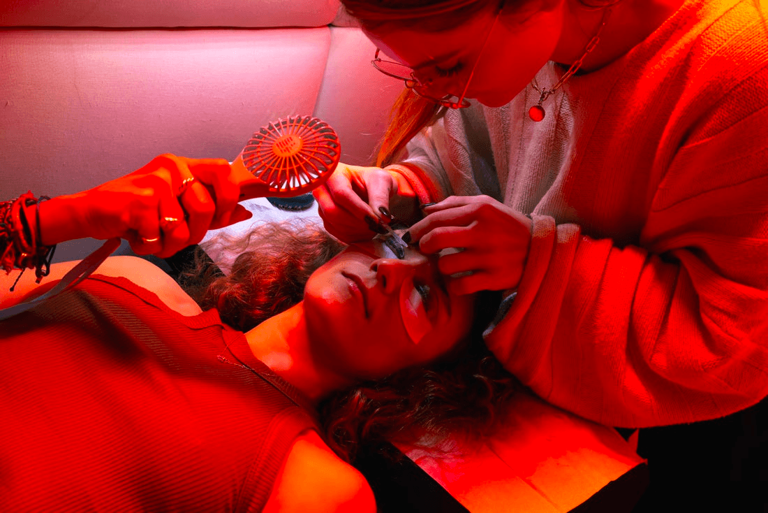 An eyelash technician applies extensions to a customer's lashes under bold red and orange lighting.