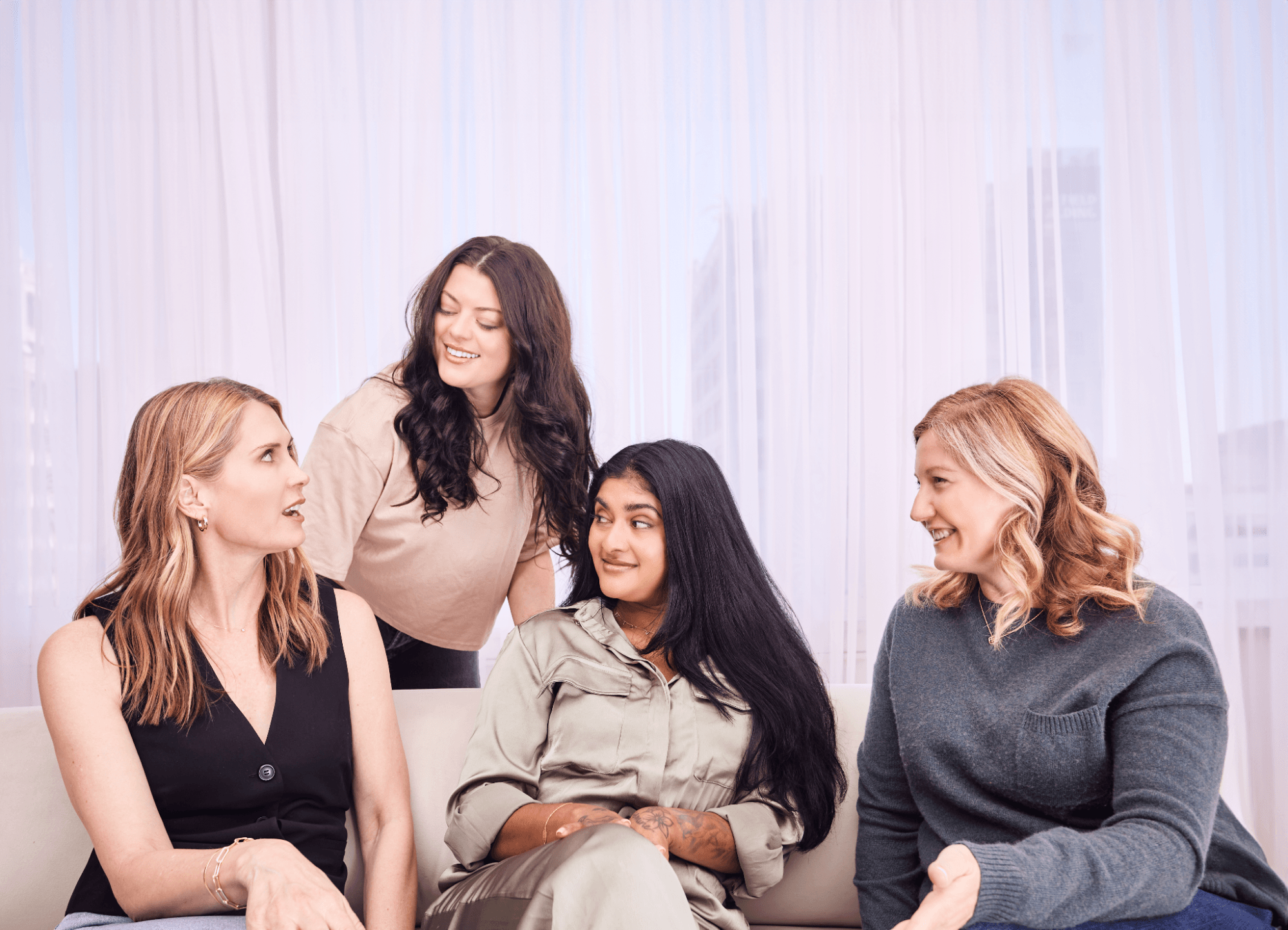 Three women on a couch, and one woman standing behind them, talk amongst themselves.