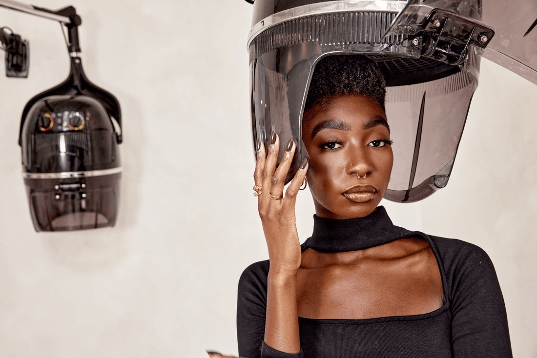 A black woman poses under a hairdrying station at a natural hair salon.