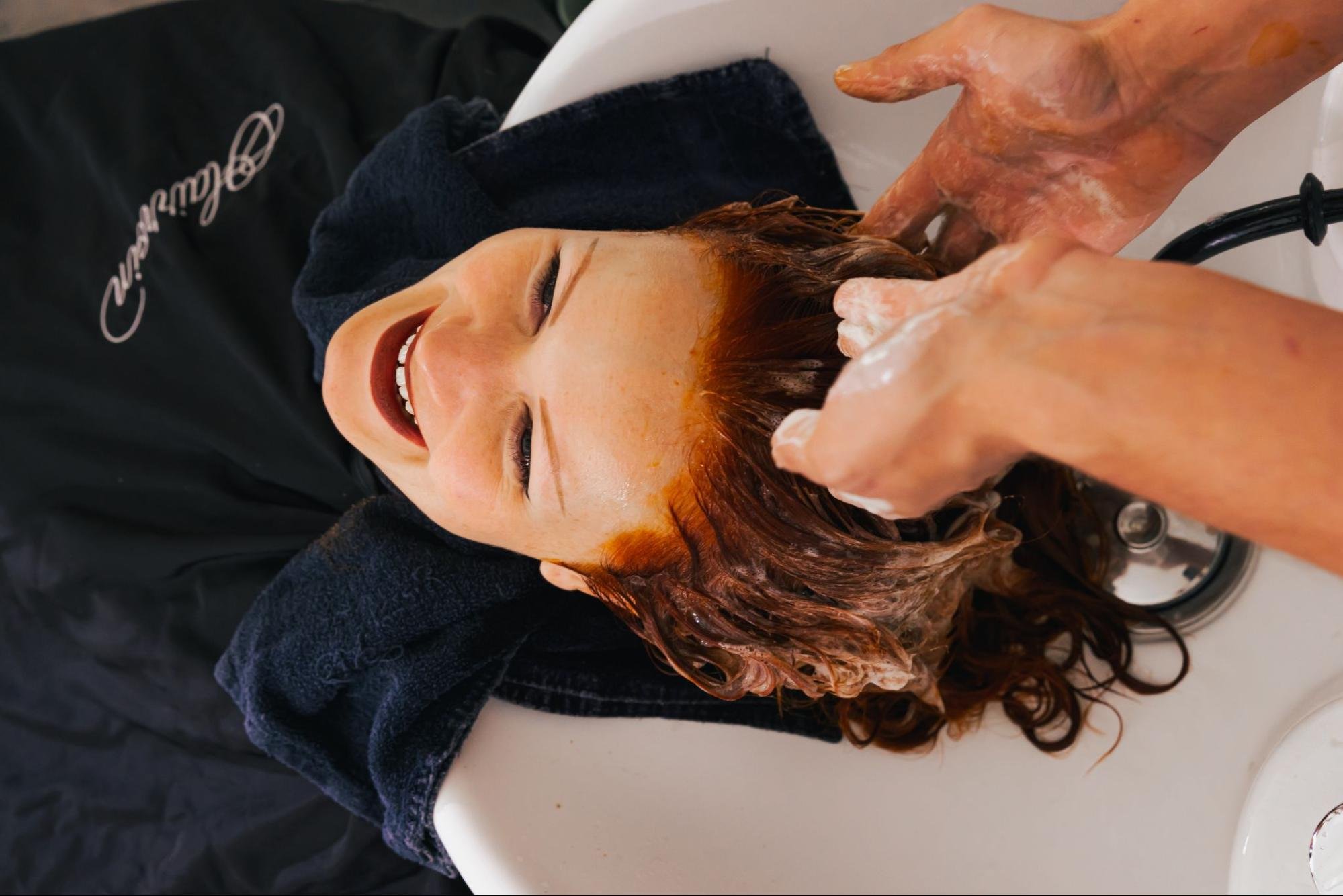 A hair salon client with bright red hair leans back over a hair wash station while a stylist washes their hair.