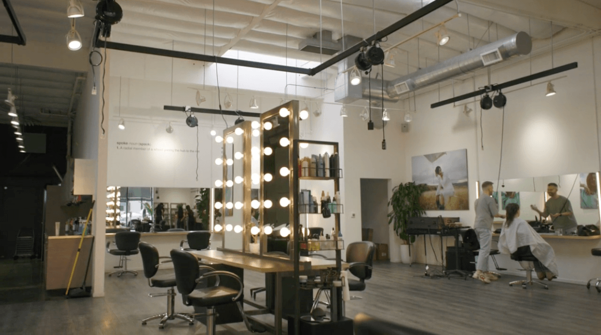 The interior of a Spoke & Weal salon. Four salon stations sit in the middle of the room, featuring tall mirrors surrounded by large backstage-inspired light bulbs. A stylist works at a station at the corner wall.
