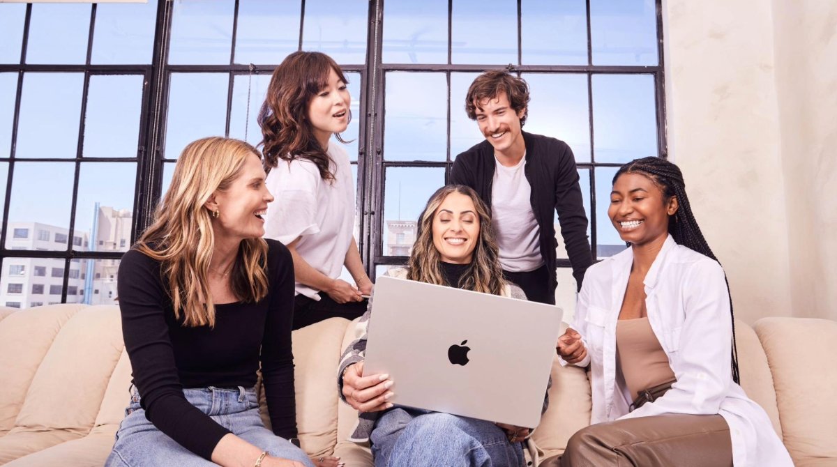 Three people sitting on a couch with two people standing behind the couch and all are looking at a laptop.