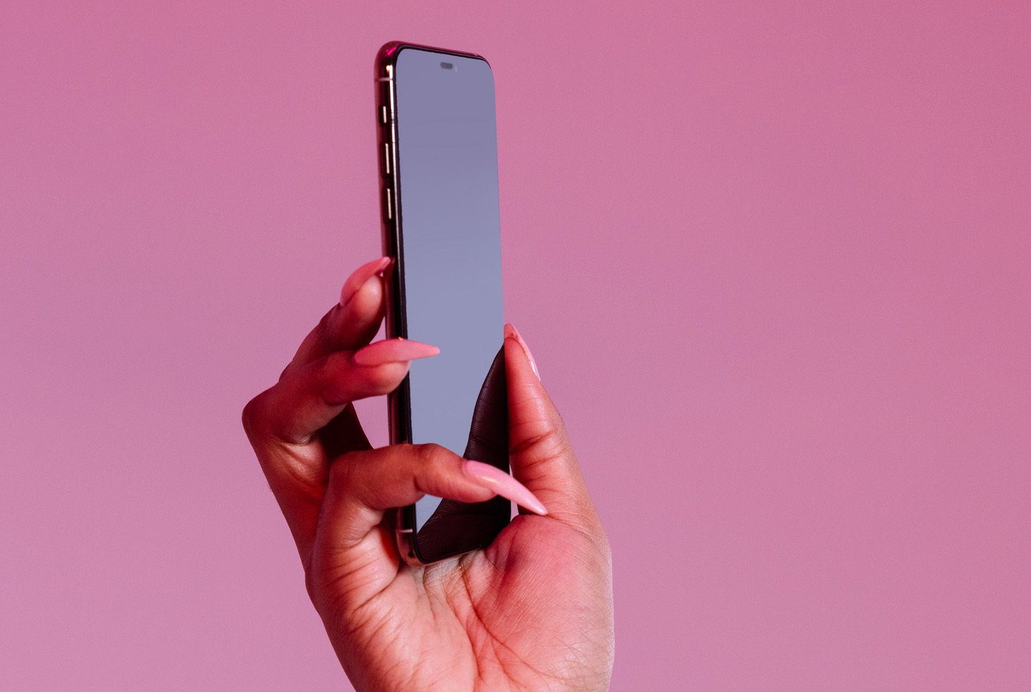 A hand emerges from out of frame, holding a smartphone in front of a pink background.