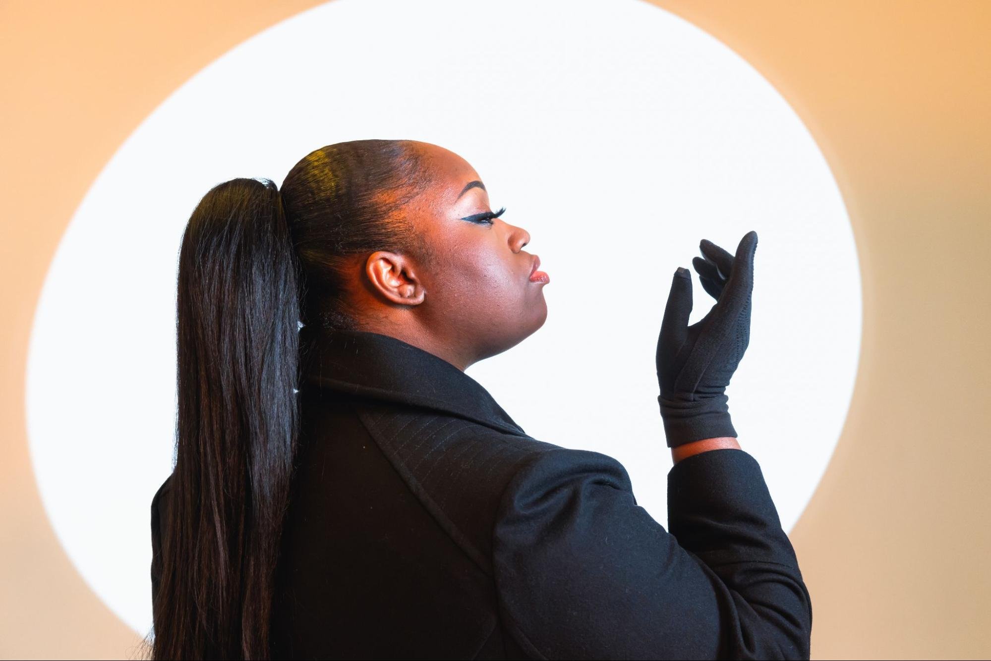 Profile view of a person with a tight ponytail framed by a bright orange background. They are holding their gloved hand in front of their face.