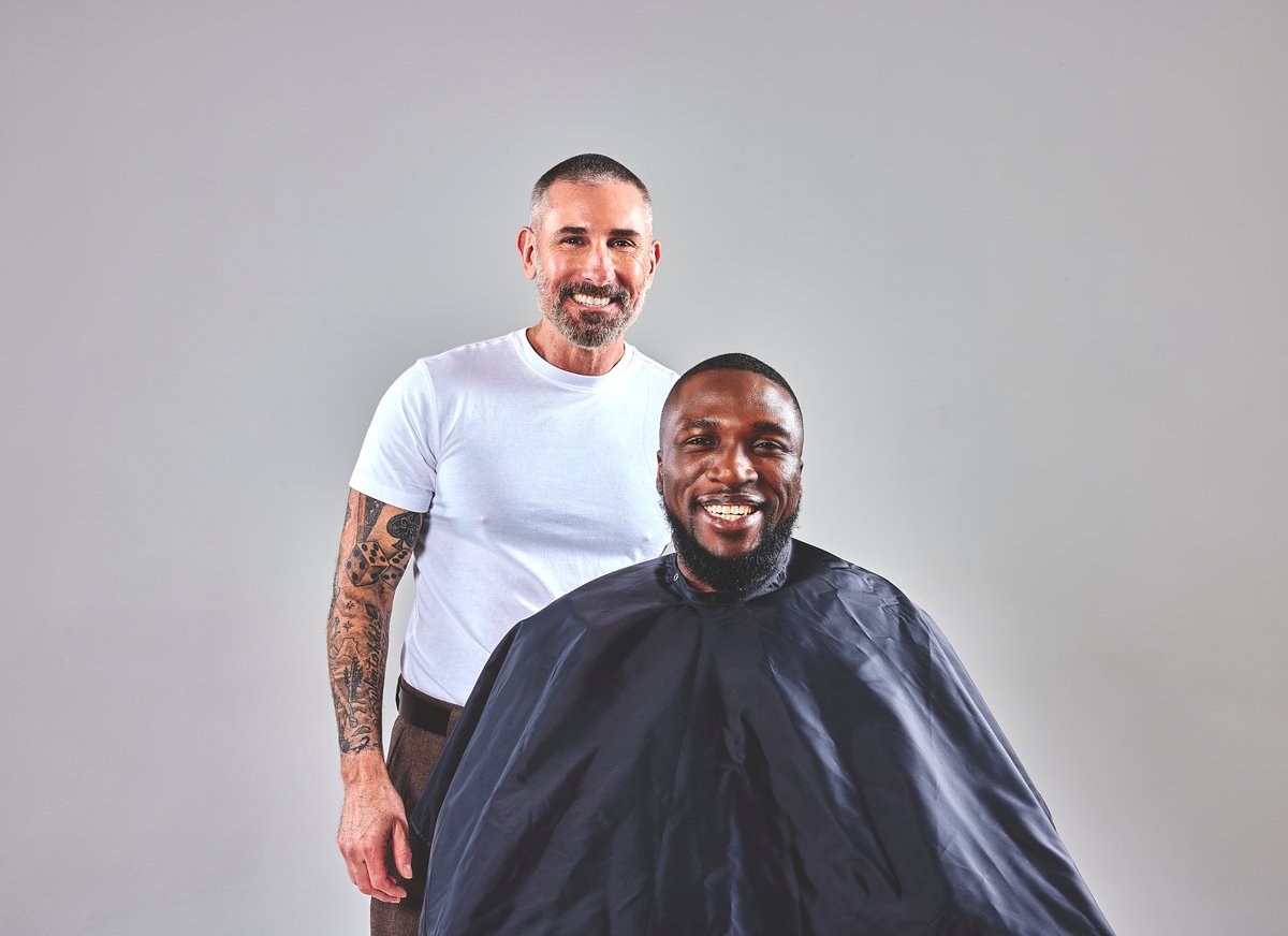 A barber services his client as they both smile at the camera.
