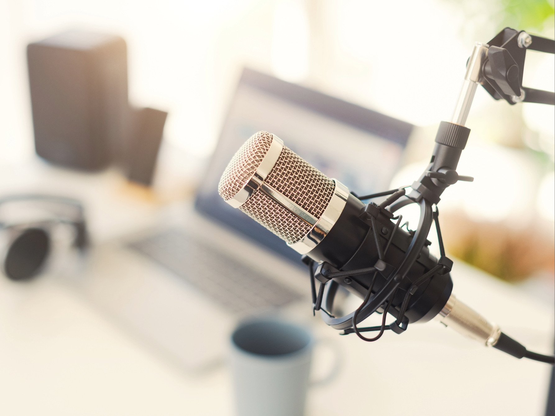A high-quality microphone hangs from its mount in the foreground, while a laptop, pair of headphones, and cup of coffee can be seen on a desk in the background.