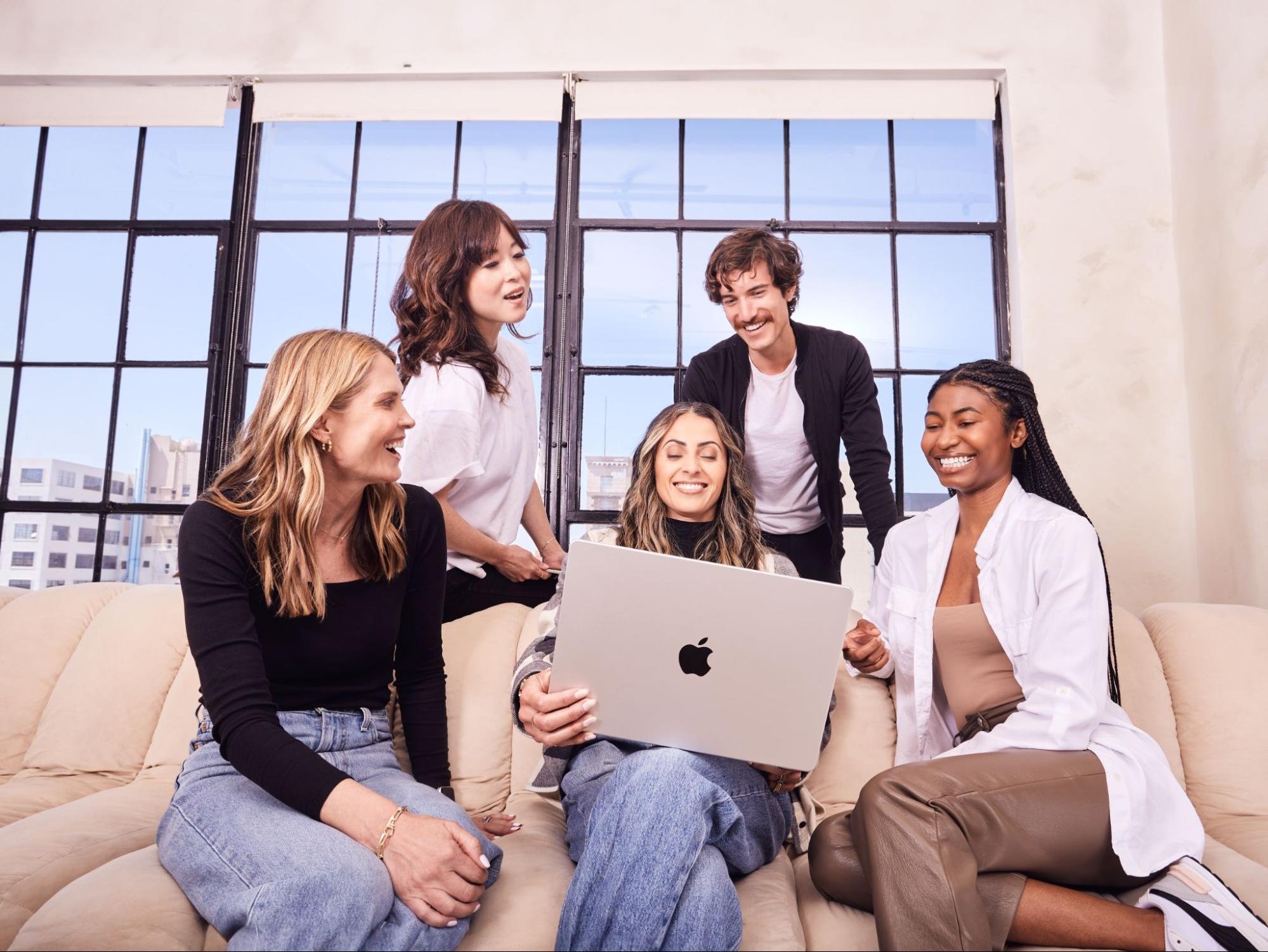 A group of five happy employees huddle around a laptop.