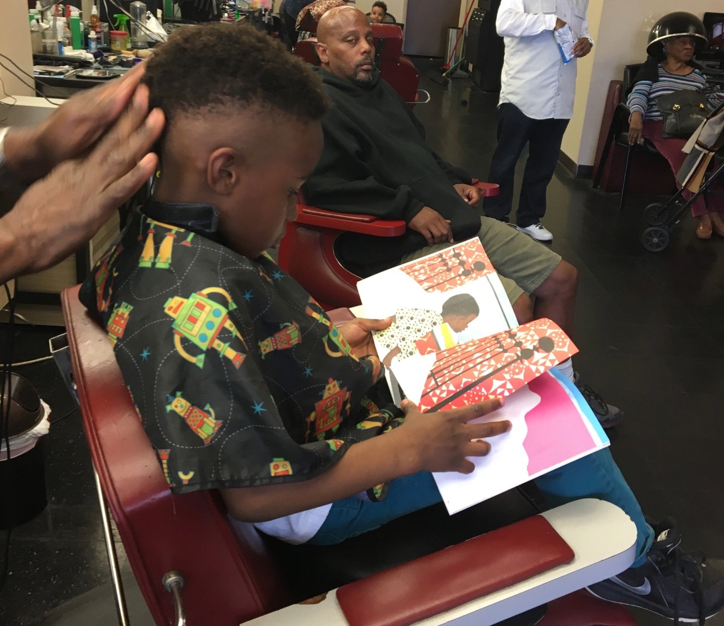 A child reads a colorful picture book while getting a haircut at a barbershop.