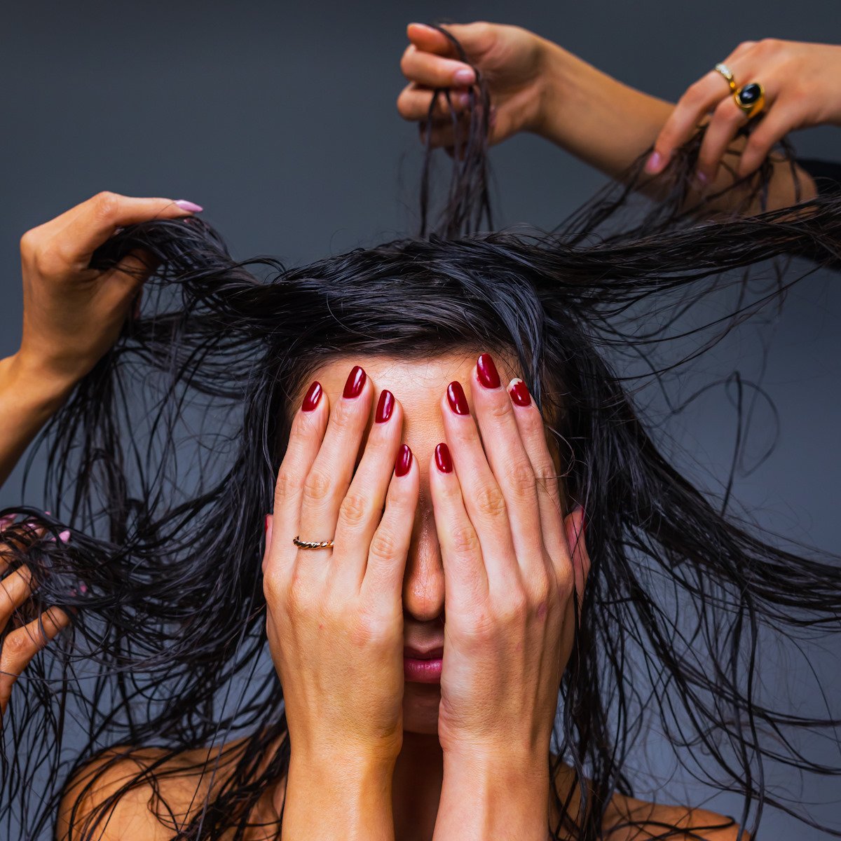 A person with long black hair covers their eyes as several hands reach in from offscreen to lift, twist, and manipulate it.