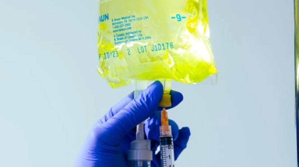 A pair of gloved hands apply a syringe to an IV bag filled with a bright yellow liquid.