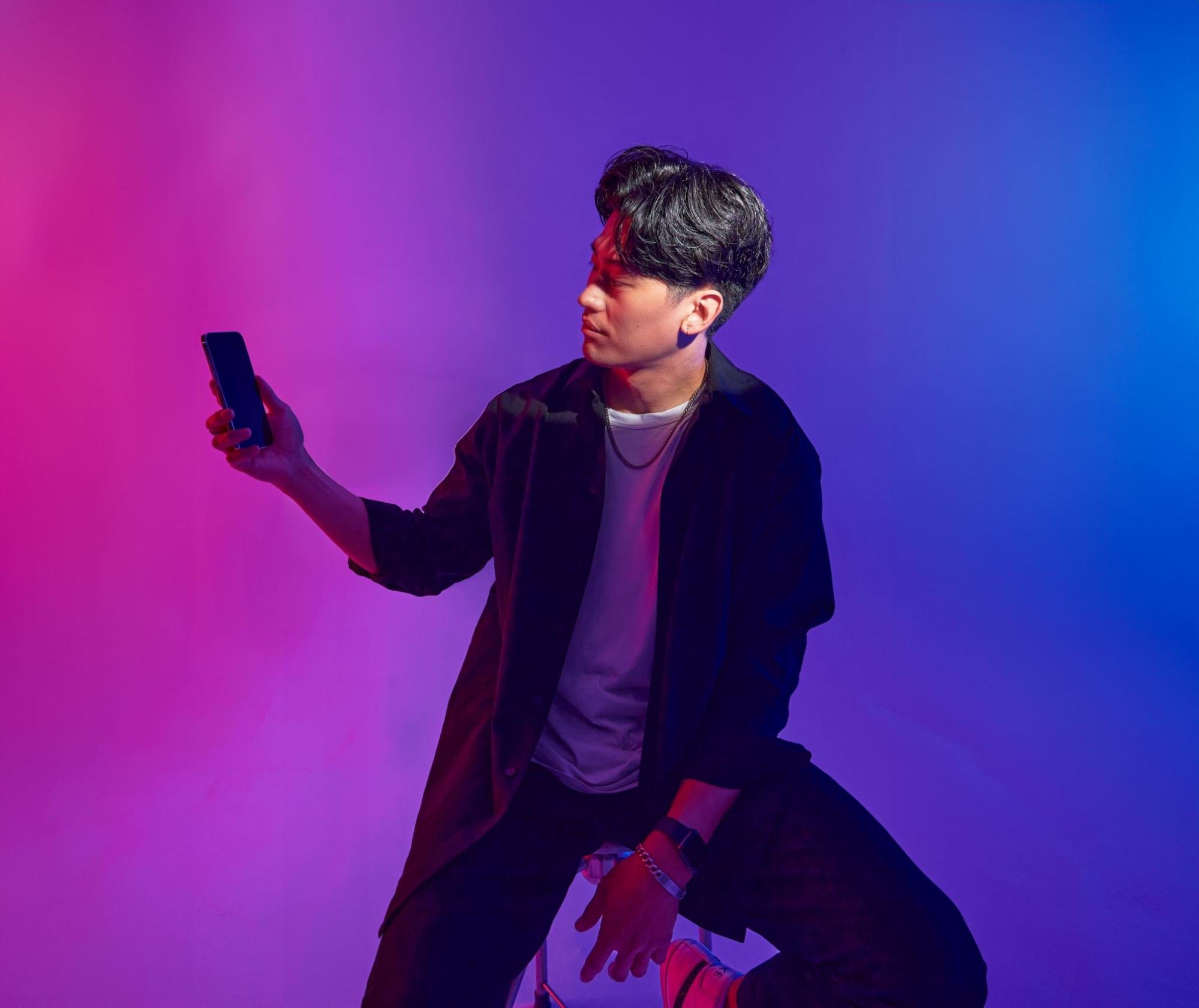 A person sits on a stool and holds a cell phone off to their side. Their face is lit by an off-screen light, setting it off from the purple and pink background.