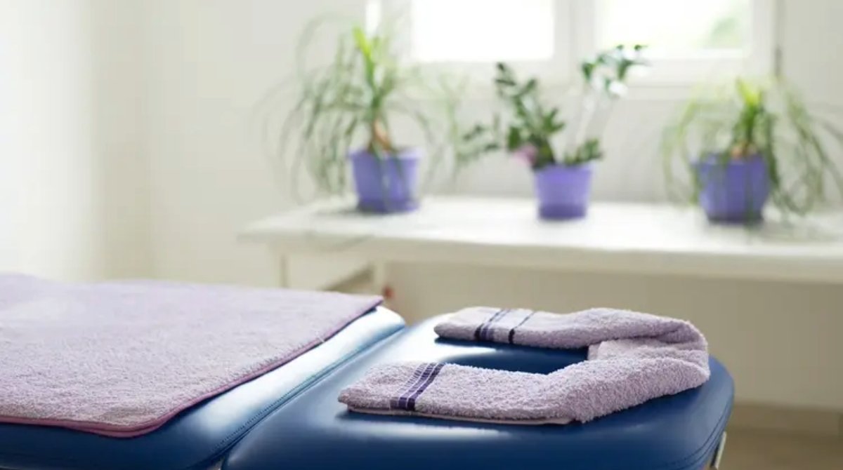 Lavender towels on a light blue massage table , with houseplants by a window in the background.