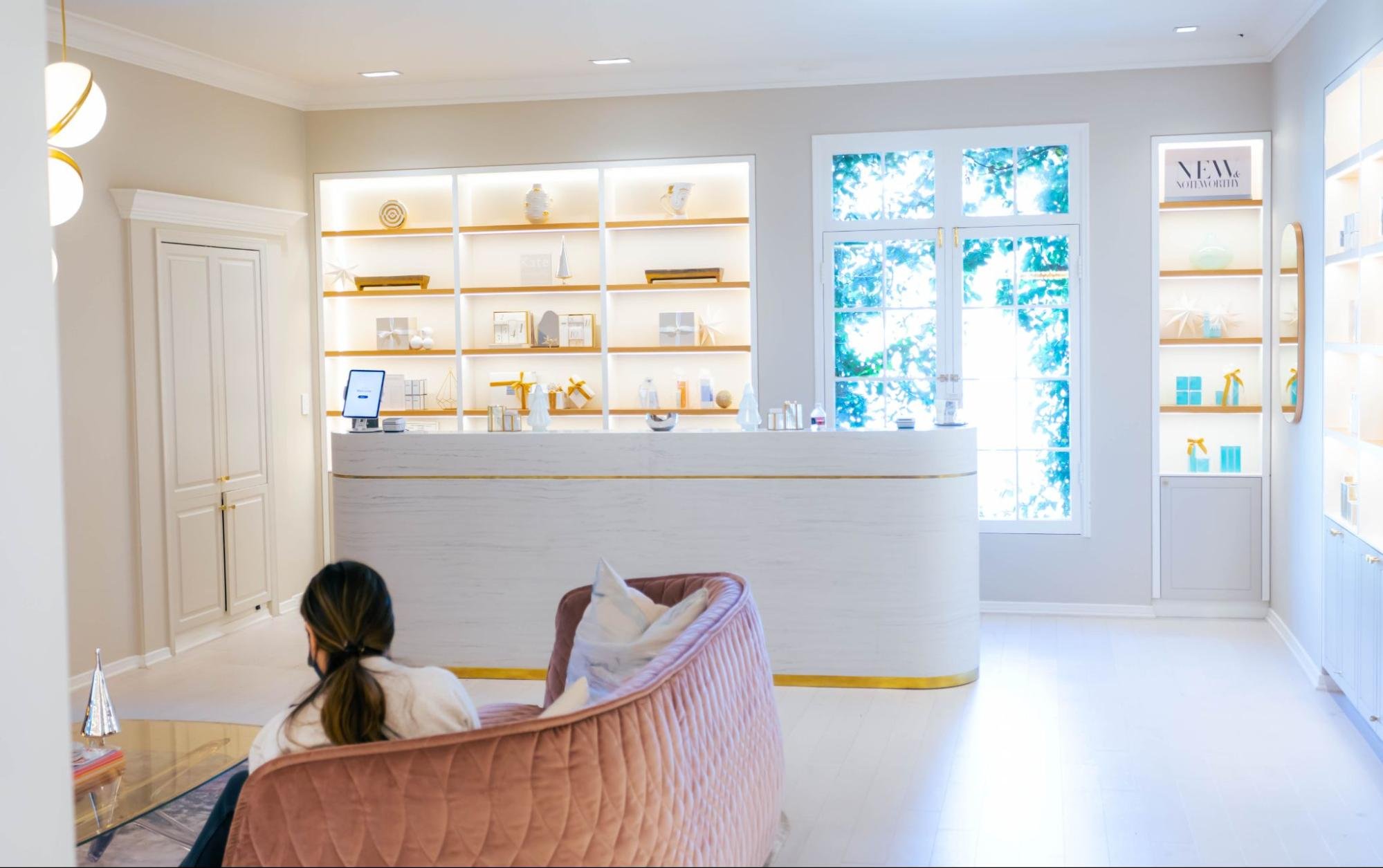 A client sits on a couch in the waiting area of a spa adorned with white walls and gold trim.