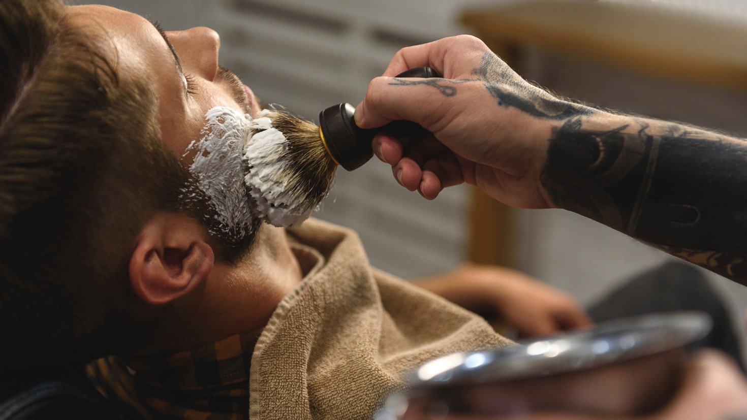 Man prepares to shave a man's face and brushes shaving cream onto his chin.