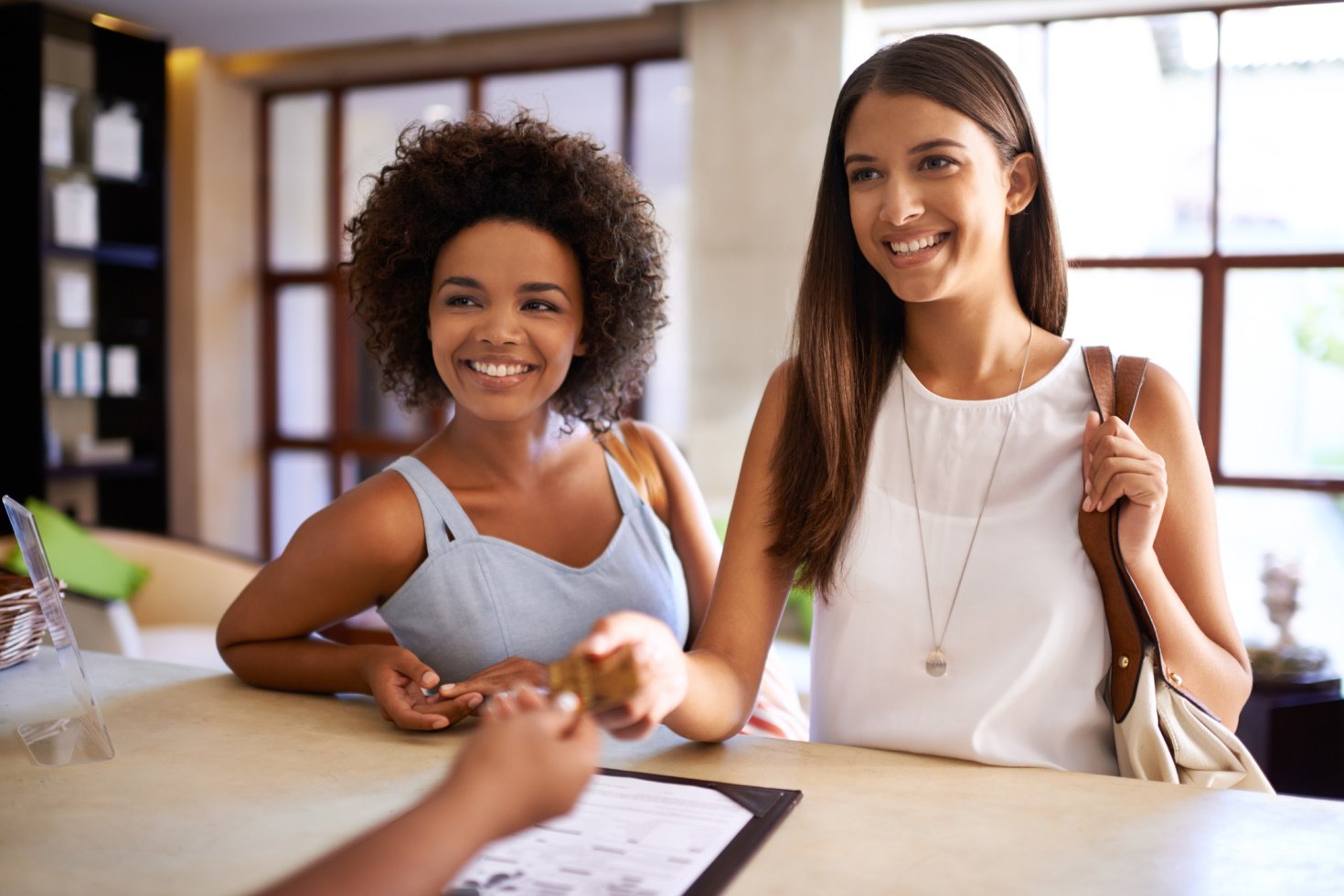 Two smiling women stand at a reception desk as one hands her payment card to the person standing opposite.