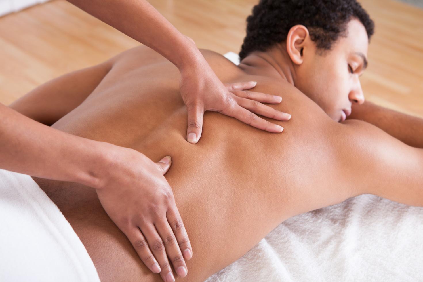 A man lies on a massage table with his eyes closed and a towel covering him from the waist down while two hands massage his back.