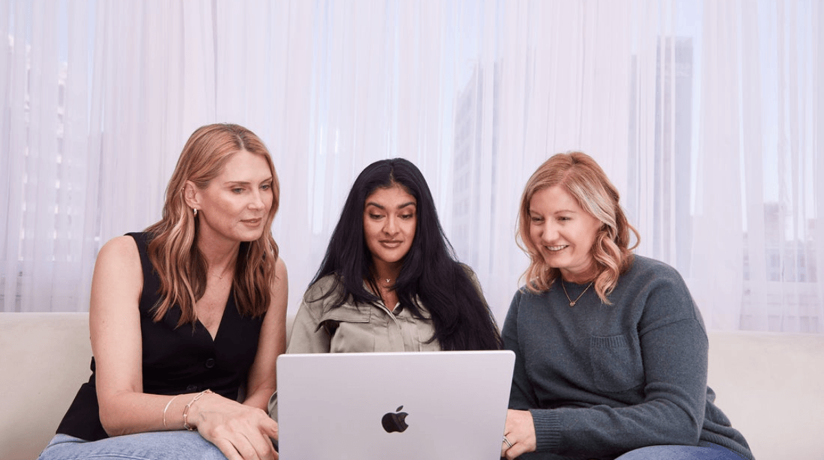Three women sitting on a couch looking at a laptop.