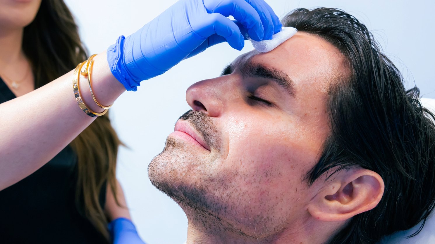 Closeup on a person after receiving an injection in their forehead. One hand in blue medical gloves holds a white cotton pad where the injection was administered.