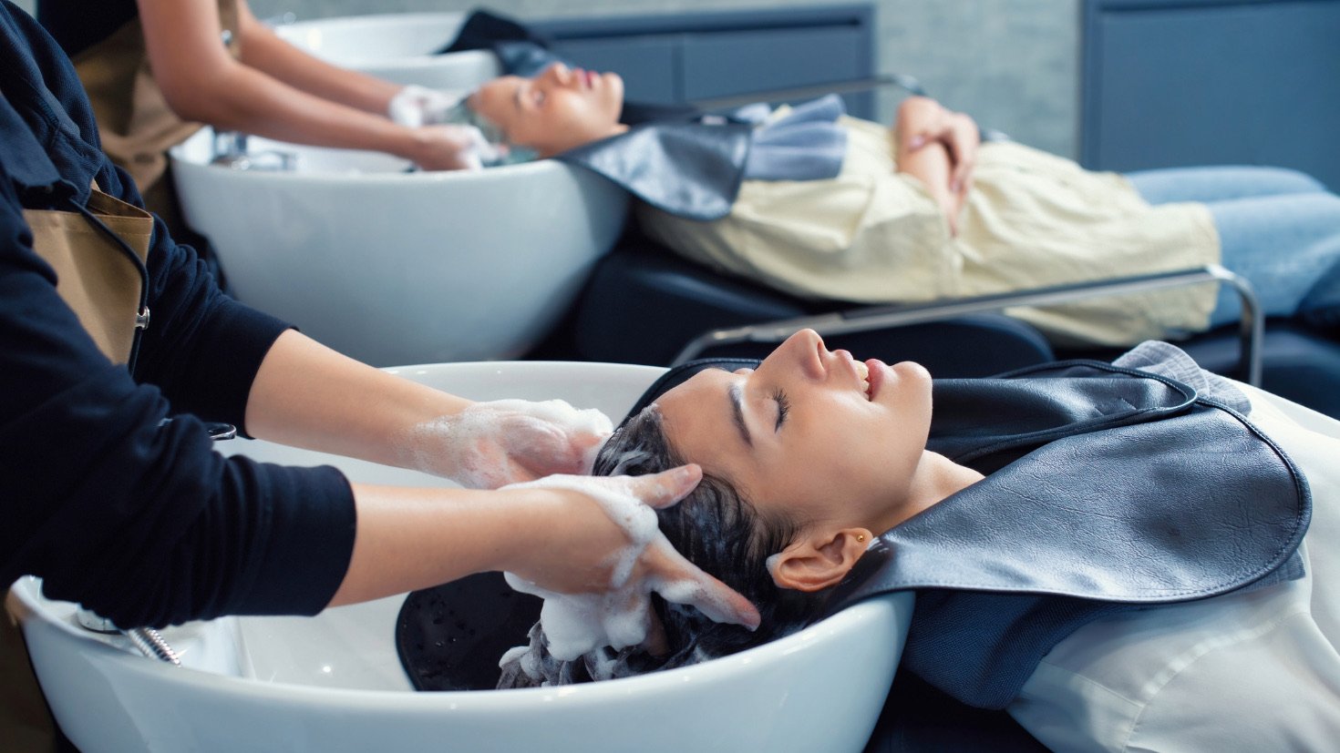 Two clients lay their heads back in adjacent sinks while stylists wash their hair.