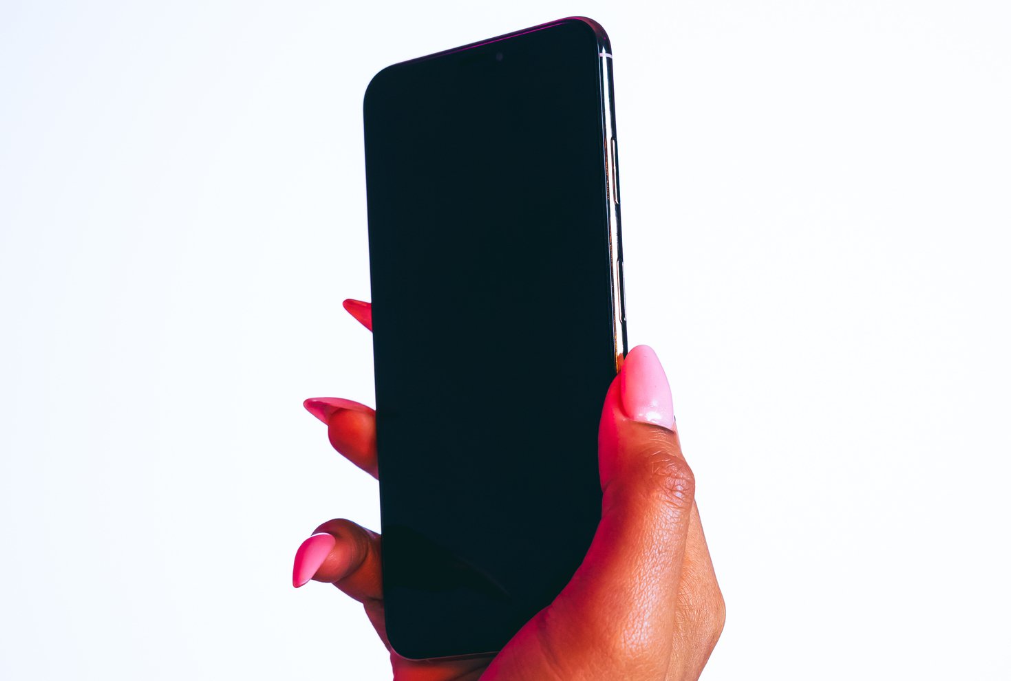 A hand with pink nails holds up a smartphone in front of a white background.