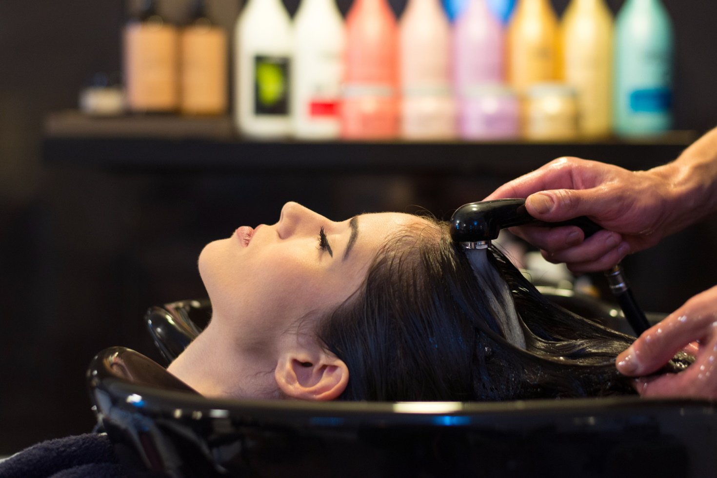 A profile view of a young woman receiving a hair rinse with colorful hair products out of focus in the background.