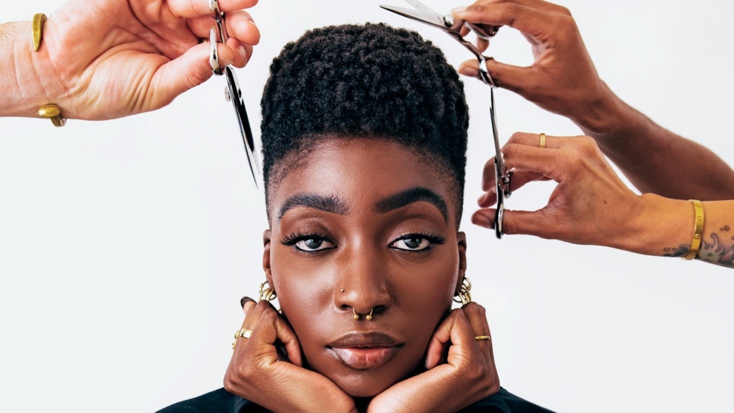 Close up of woman with buzzed hair looking straight into the lens as 3 hands each with a pair of scissors float just above her head.