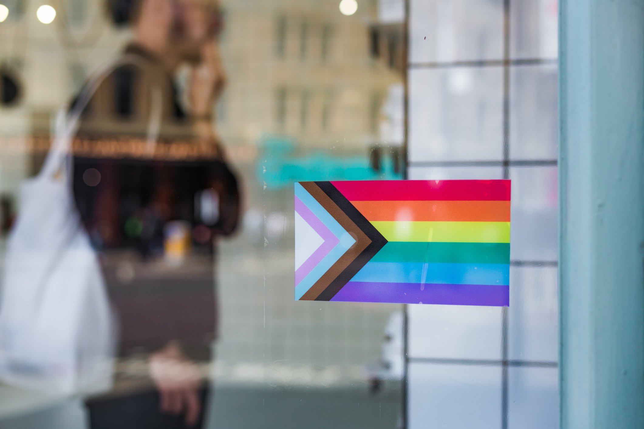 A Progress Pride flag sticker is in a shop window in the foreground. A customer can be seen in the background out of focus.
