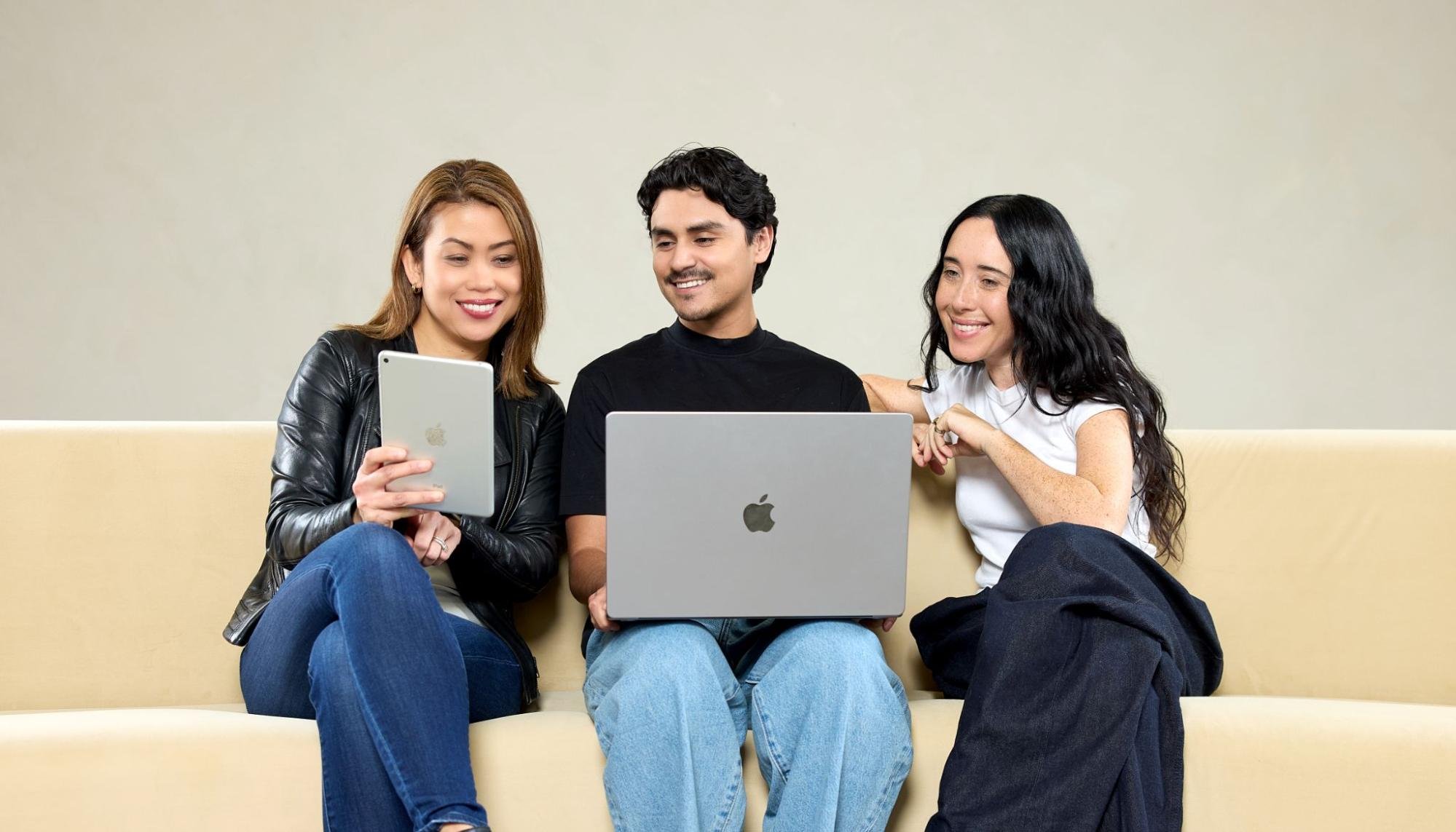 Three people sitting on a couch, smiling as they look at a laptop screen and tablet.