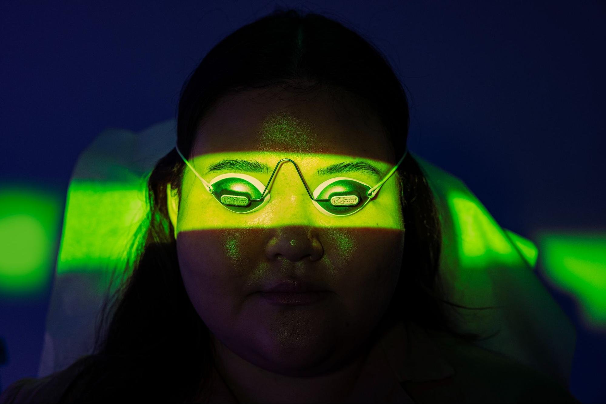 A medspa client wears protective eye coverings as they undergo a skin treatment, a beam of green light passing over their eyes.